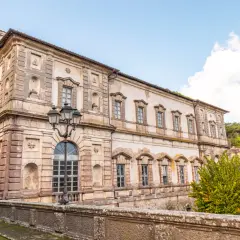 Il **Palazzo Chigi Soriano nel Cimino**, con la sua facciata imponente e le finestre eleganti, si staglia contro un cielo azzurro con nuvole bianche.
