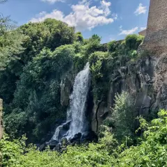 Cascata rigogliosa nel **lago di Nepi**, con rocce antiche e vegetazione lussureggiante sotto un cielo azzurro e nuvoloso.