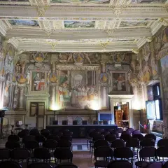 Sala affrescata nel **Palazzo dei Priori**, con sedie disposte per un evento. Soffitto e pareti riccamente decorati con scene storiche e ritratti.