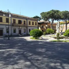 Piazza principale di Monte Romano, Viterbo, con il palazzo comunale, una fontana e alberi rigogliosi sotto un cielo azzurro.