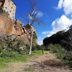 Sentiero nei pressi delle forre di Corchiano, con un edificio arroccato sulla rupe e un albero spoglio in primo piano.