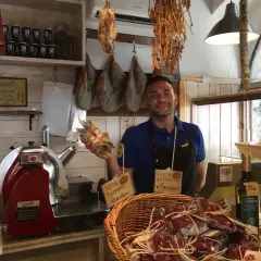 Un salumiere sorridente mostra salumi in un negozio di Bolsena. Prosciutto appeso, affettatrice e prodotti tipici invitano a scoprire i sapori locali.