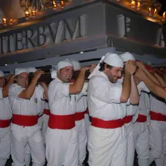Uomini con fasce rosse e copricapi bianchi trasportano un pesante manufatto illuminato da candele. La scritta "VITERBVM" è visibile. La macchina di Santa Rosa caduta è un evento drammatico.