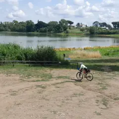 Ciclista pedala vicino al lago di Monterosi, con vegetazione rigogliosa e cielo nuvoloso.