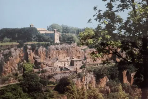 Antico complesso monastico di Santa Maria ad Rupes, incastonato nella roccia, con edifici in pietra e vegetazione rigogliosa.