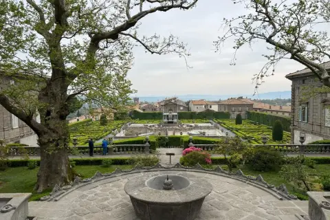 Giardino all'italiana con fontane e siepi geometriche, con un grande albero in primo piano. La serpara domina il paesaggio.
