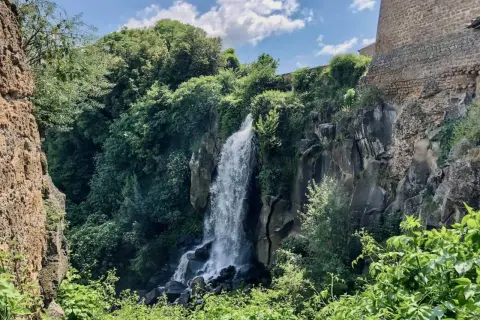 Cascata rigogliosa nel **lago di Nepi**, con rocce antiche e vegetazione lussureggiante sotto un cielo azzurro e nuvoloso.