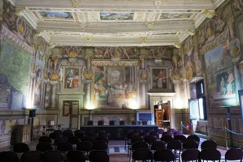 Sala affrescata nel **Palazzo dei Priori**, con sedie disposte per un evento. Soffitto e pareti riccamente decorati con scene storiche e ritratti.