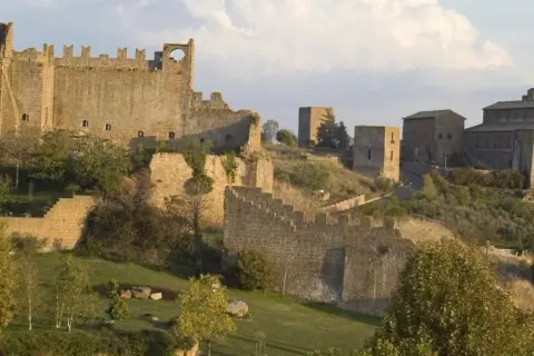 Antiche mura e torri di Tuscania VT si stagliano su una collina verdeggiante, con una chiesa imponente in lontananza.