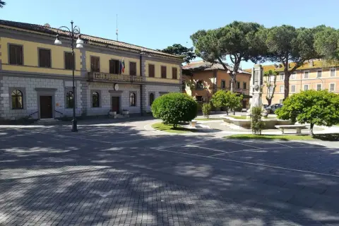 Piazza principale di Monte Romano, Viterbo, con il palazzo comunale, una fontana e alberi rigogliosi sotto un cielo azzurro.