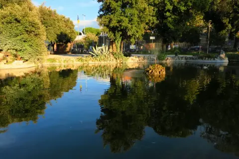 Un laghetto con riflessi verdi e azzurri, un'isola rocciosa con una fontana e alberi rigogliosi in un prato giardino a Viterbo.