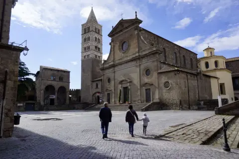 Una famiglia passeggia nella storica piazza San Lorenzo a Viterbo, ammirando la maestosa cattedrale e il suo campanile.