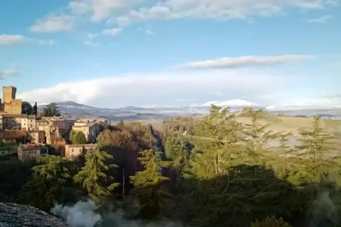 Borgo medievale con torre imponente, immerso nel verde del comune di Proceno, con colline nebbiose all'orizzonte.