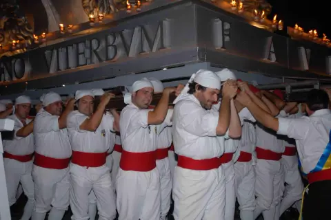 Uomini con fasce rosse e copricapi bianchi trasportano un pesante manufatto illuminato da candele. La scritta "VITERBVM" è visibile. La macchina di Santa Rosa caduta è un evento drammatico.
