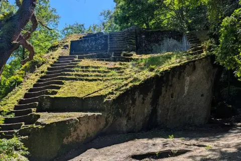 Antica piramide etrusca scavata nella roccia, ricoperta di muschio e circondata da alberi secolari.