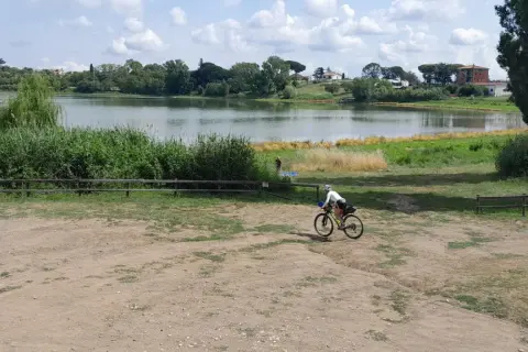Ciclista pedala vicino al lago di Monterosi, con vegetazione rigogliosa e cielo nuvoloso.