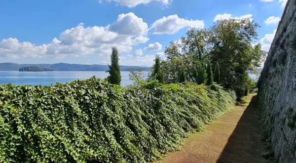 Un sentiero fiancheggiato da un muro di pietra e una siepe rigogliosa conduce alla vista del **lago di Capodimonte**, con un'isola in lontananza sotto un cielo azzurro e nuvoloso.