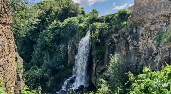 Cascata rigogliosa nel **lago di Nepi**, con rocce antiche e vegetazione lussureggiante sotto un cielo azzurro e nuvoloso.