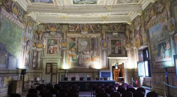 Sala affrescata nel **Palazzo dei Priori**, con sedie disposte per un evento. Soffitto e pareti riccamente decorati con scene storiche e ritratti.