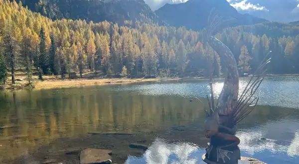 Scultura di un pesce con canna da pesca sul **lago di San Pellegrino**, circondato da boschi autunnali e montagne.