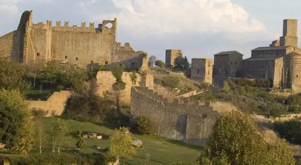 Antiche mura e torri di Tuscania VT si stagliano su una collina verdeggiante, con una chiesa imponente in lontananza.