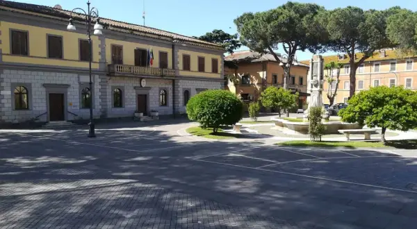 Piazza principale di Monte Romano, Viterbo, con il palazzo comunale, una fontana e alberi rigogliosi sotto un cielo azzurro.