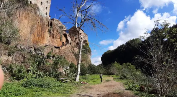 Sentiero nei pressi delle forre di Corchiano, con un edificio arroccato sulla rupe e un albero spoglio in primo piano.