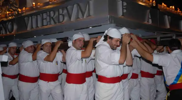 Uomini con fasce rosse e copricapi bianchi trasportano un pesante manufatto illuminato da candele. La scritta "VITERBVM" è visibile. La macchina di Santa Rosa caduta è un evento drammatico.