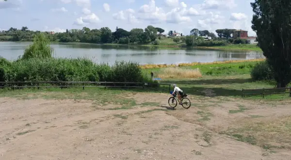 Ciclista pedala vicino al lago di Monterosi, con vegetazione rigogliosa e cielo nuvoloso.