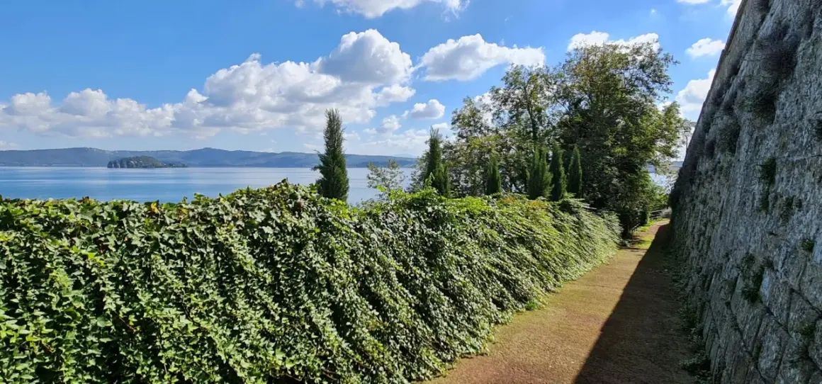 Un sentiero fiancheggiato da un muro di pietra e una siepe rigogliosa conduce alla vista del **lago di Capodimonte**, con un'isola in lontananza sotto un cielo azzurro e nuvoloso.