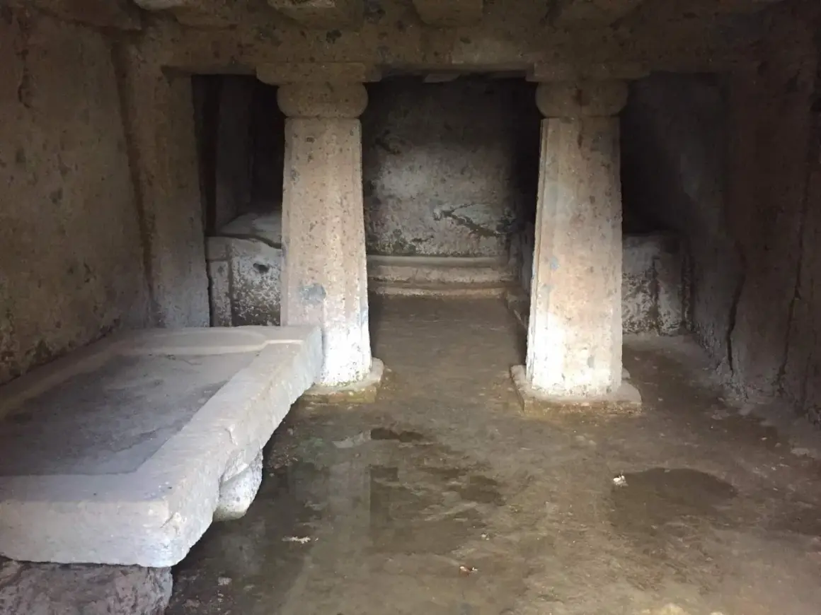 Interno di una tomba a dado scavata nella roccia, con un sarcofago in pietra e colonne grezze.