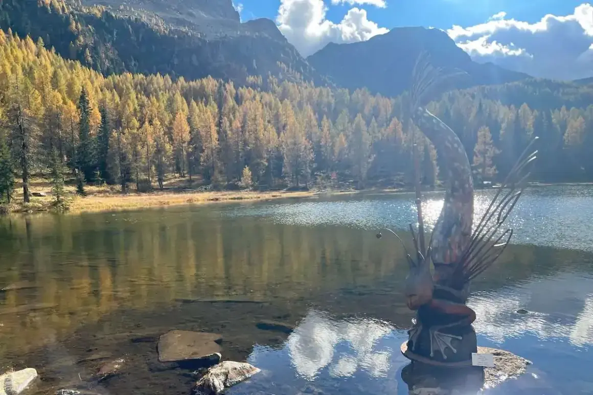 Scultura di un pesce con canna da pesca sul **lago di San Pellegrino**, circondato da boschi autunnali e montagne.