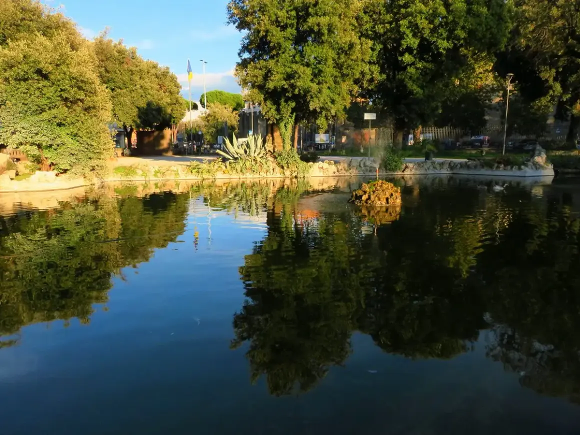 Un laghetto con riflessi verdi e azzurri, un'isola rocciosa con una fontana e alberi rigogliosi in un prato giardino a Viterbo.