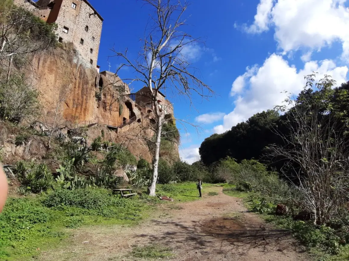 Sentiero nei pressi delle forre di Corchiano, con un edificio arroccato sulla rupe e un albero spoglio in primo piano.