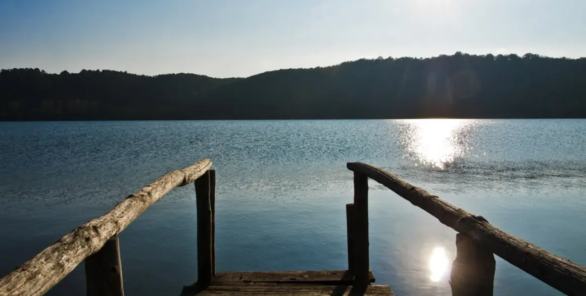 Un pontile in legno si affaccia sul lago di Mezzano, con il sole che brilla sull'acqua e le colline sullo sfondo.