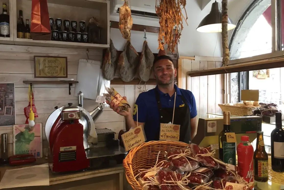 Un salumiere sorridente mostra salumi in un negozio di Bolsena. Prosciutto appeso, affettatrice e prodotti tipici invitano a scoprire i sapori locali.
