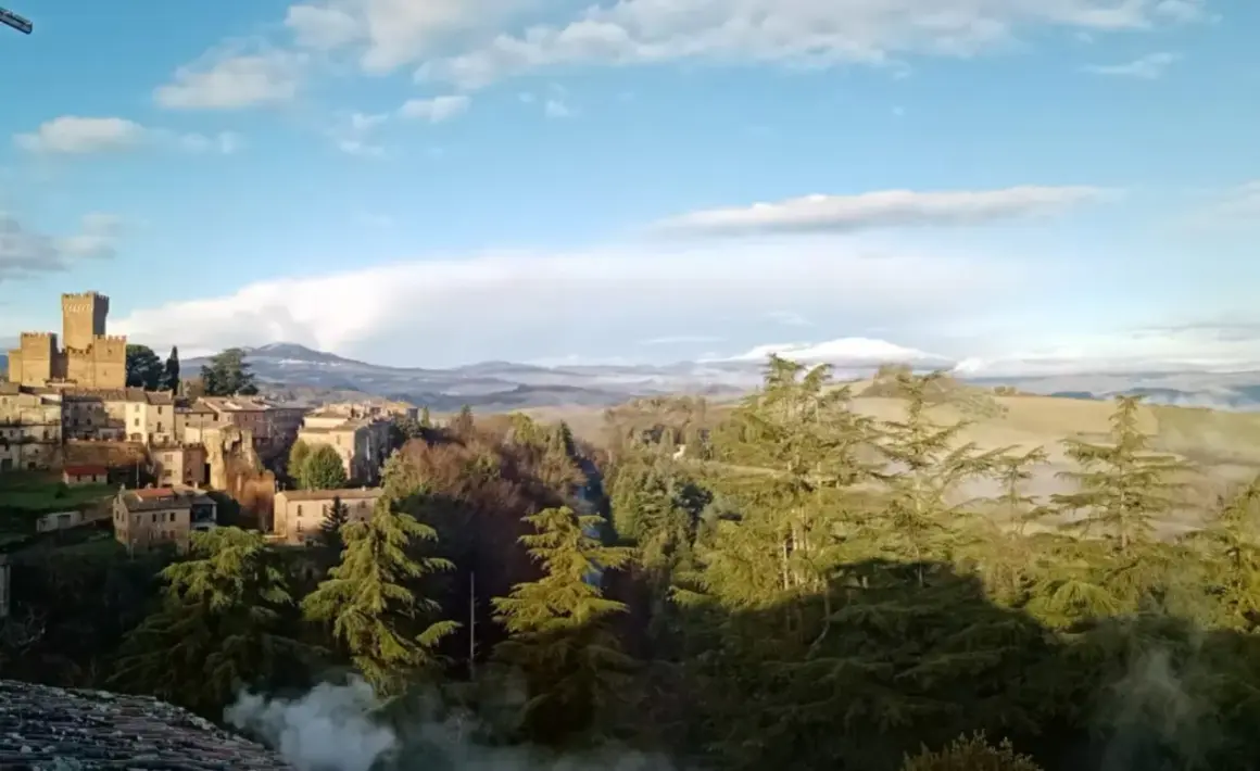 Borgo medievale con torre imponente, immerso nel verde del comune di Proceno, con colline nebbiose all'orizzonte.