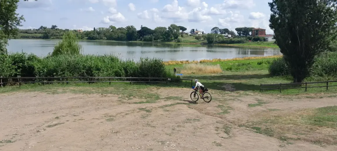 Ciclista pedala vicino al lago di Monterosi, con vegetazione rigogliosa e cielo nuvoloso.