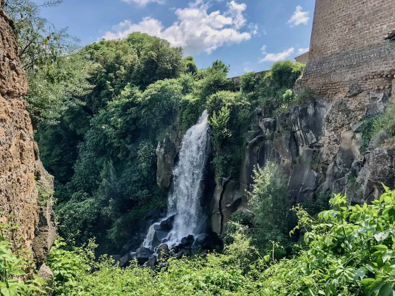 Cascata rigogliosa e rocciosa, con vegetazione lussureggiante che incornicia la sorgente Nepi. Un muro antico sovrasta il paesaggio.