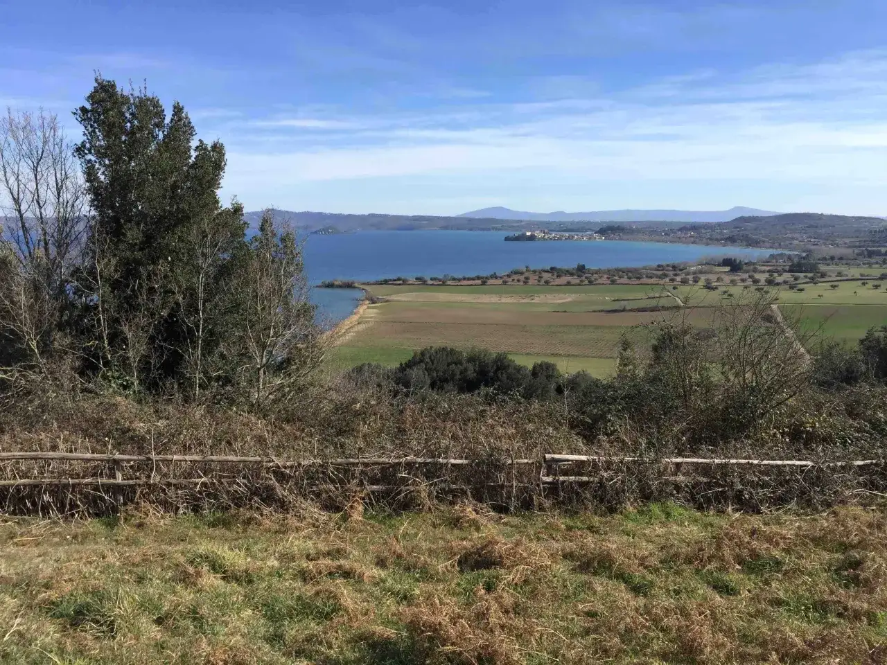 Vista panoramica dal monte Bisenzio: lago azzurro, campi verdi e un borgo in lontananza sotto un cielo limpido.