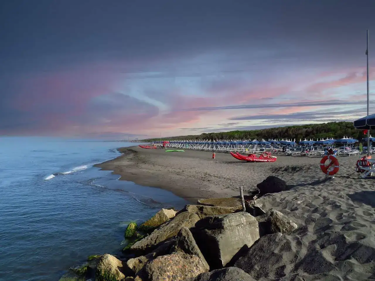 Spiaggia di Montalto Mare al tramonto, con ombrelloni, lettini, una barca di salvataggio rossa e il mare calmo sotto un cielo colorato.