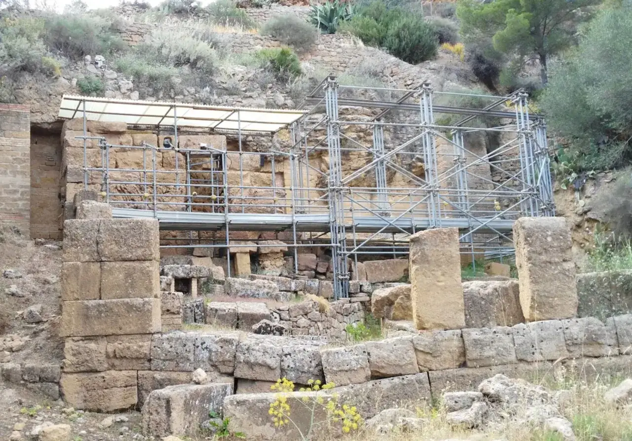 Resti archeologici di Santa Maria ad Rupes con impalcature metalliche e tettoia protettiva. Blocchi di pietra grezza e vegetazione circostante.