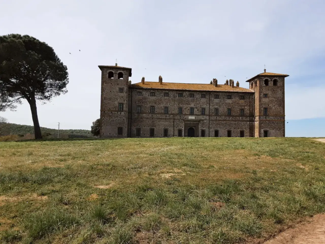 Antico palazzo fortificato a Monte Romano, Viterbo, con torri angolari e tetto spiovente arancione, circondato da un prato verde.