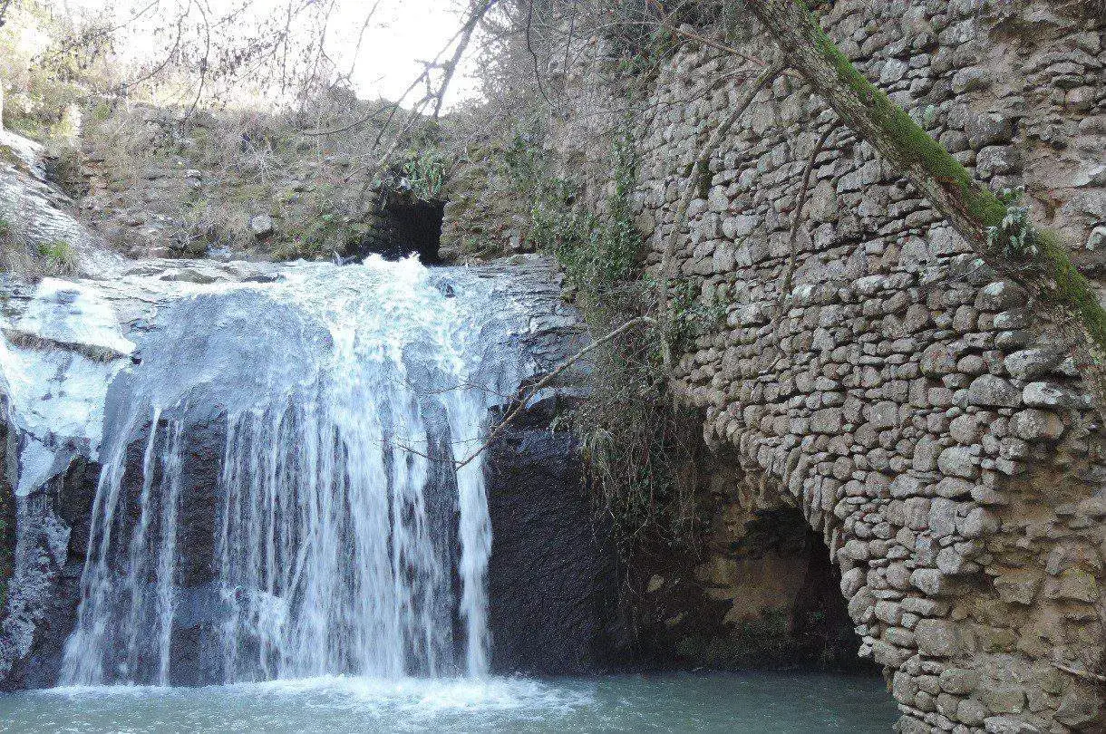 Cascata nelle forre di Corchiano, con un antico muro in pietra che forma un arco sopra l'acqua.