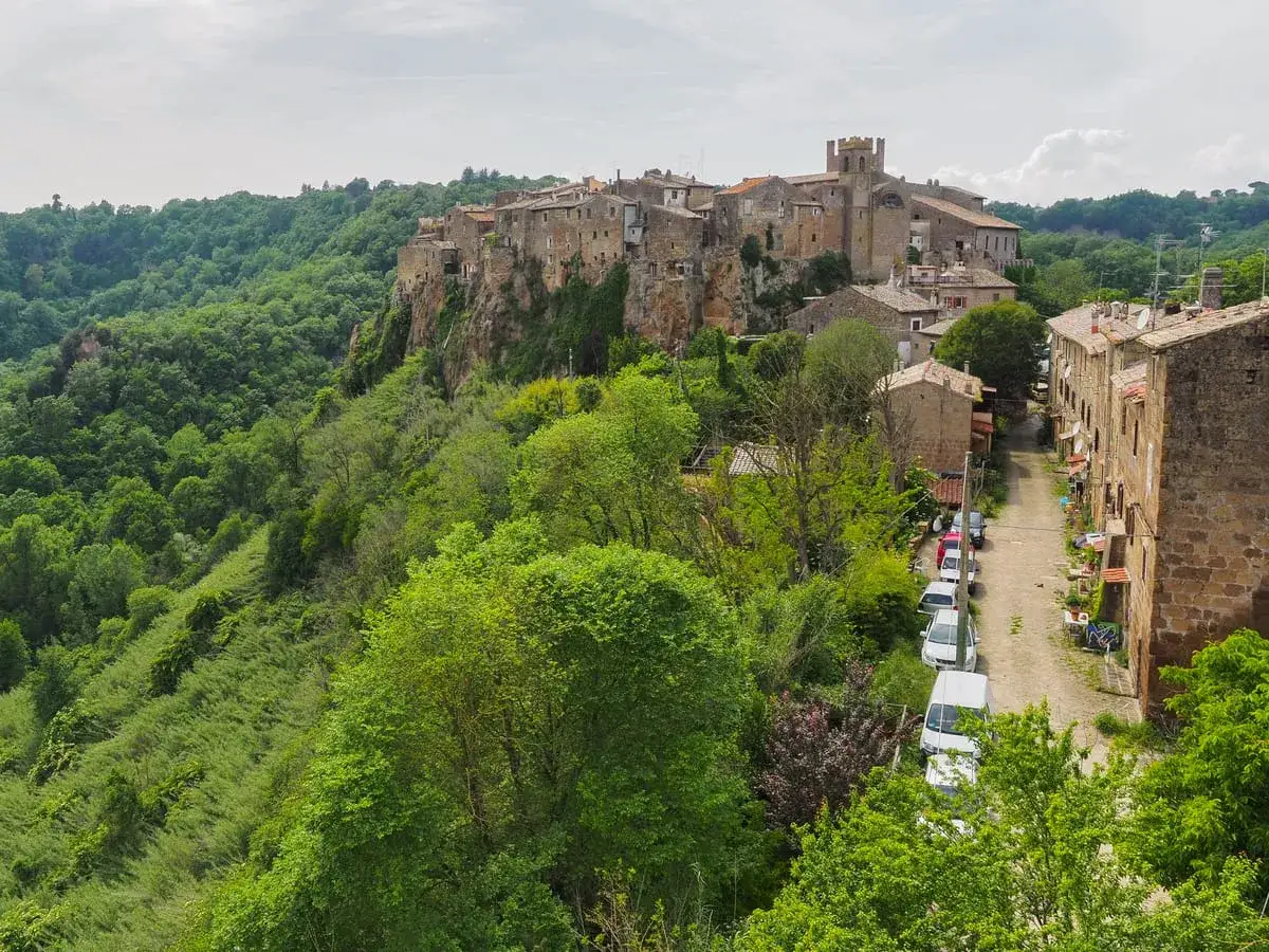 Borgo medievale arroccato su una rupe, circondato da una rigogliosa vegetazione. Un vicolo con auto parcheggiate attraversa il paese, offrendo uno scorcio di Calcata e dintorni.