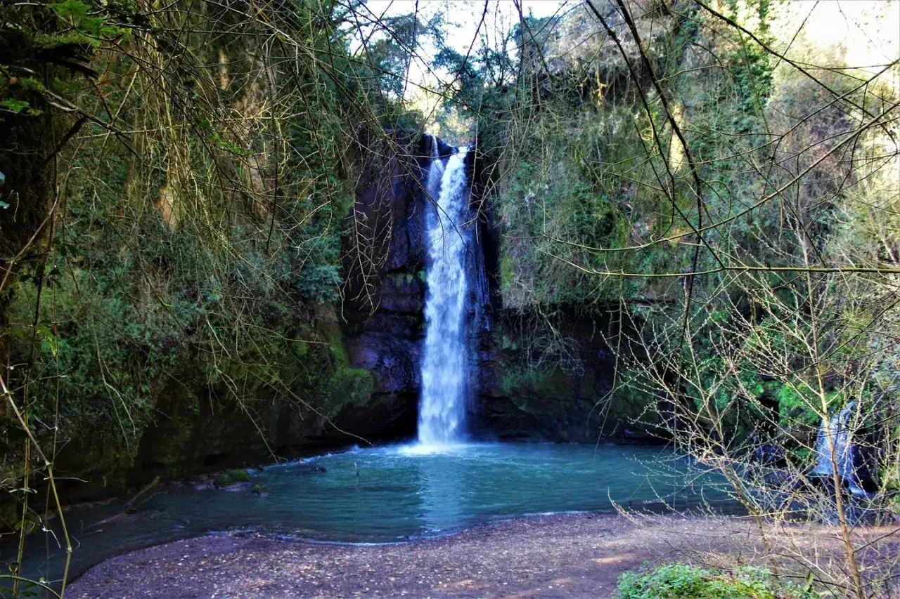 La cascata del picchio scende fragorosa in una pozza azzurra, incorniciata da rocce muschiose e alberi spogli.