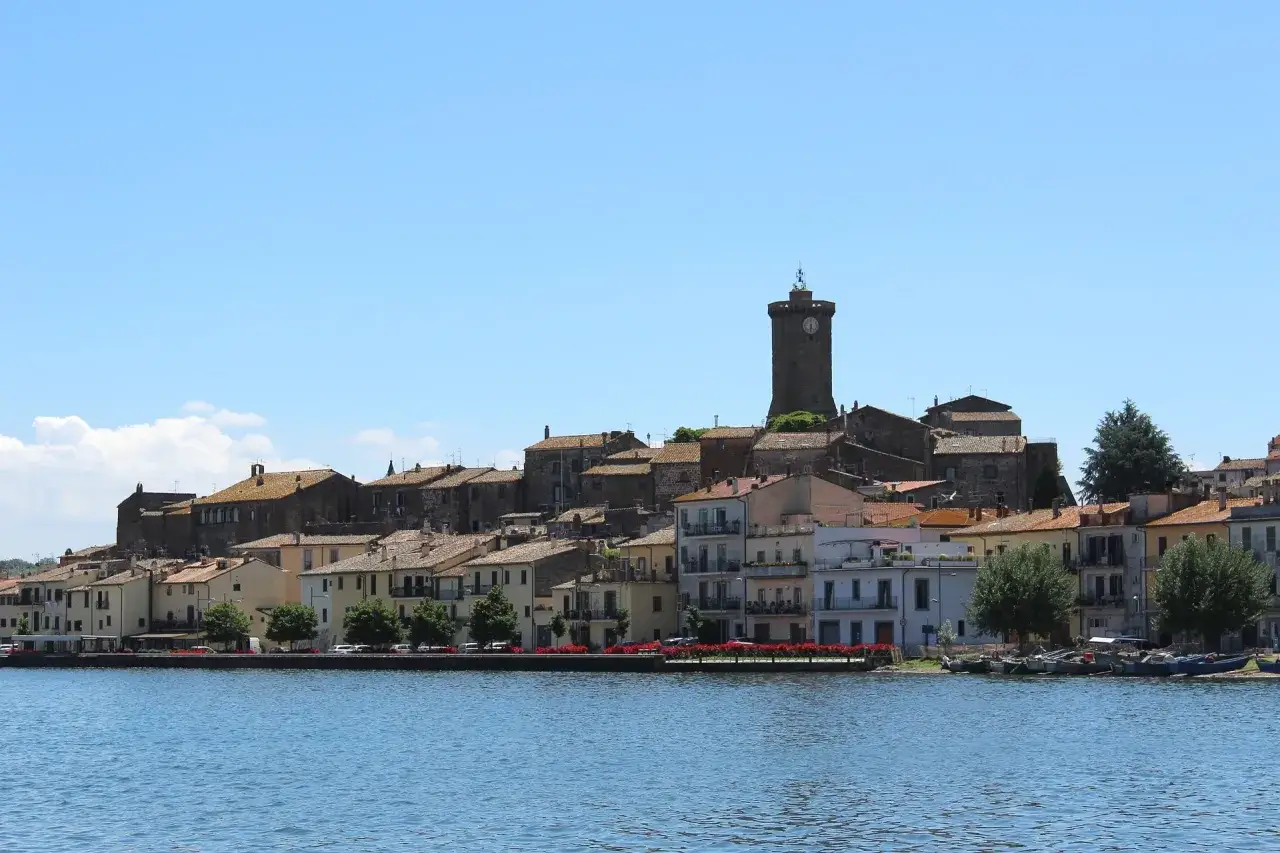 Vista del borgo di Isola Martana con le sue case colorate e la torre dell'orologio, affacciato su un lago tranquillo.