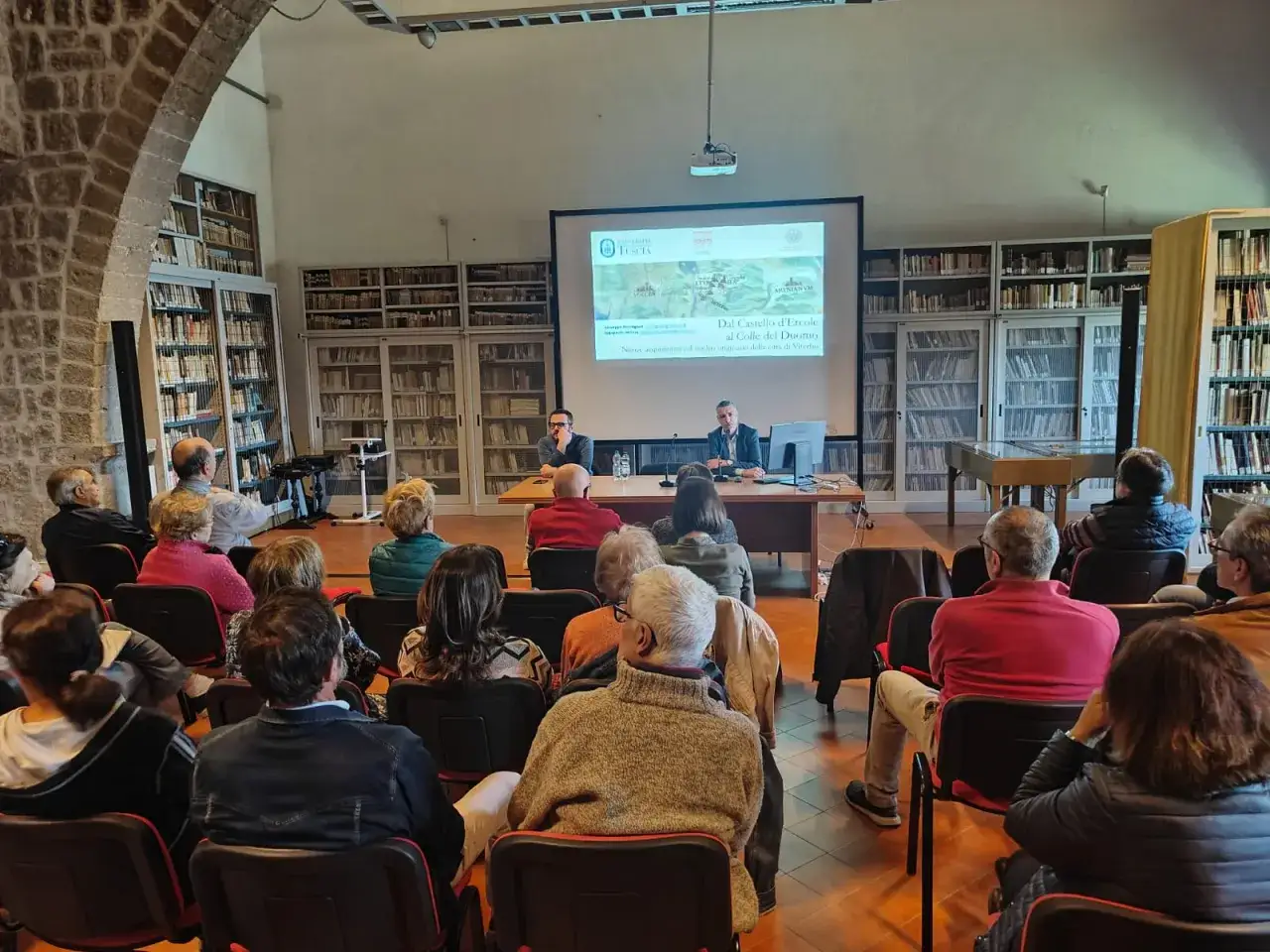 Conferenza sul Duomo di Viterbo, con relatori e pubblico in sala, circondati da scaffali di libri.