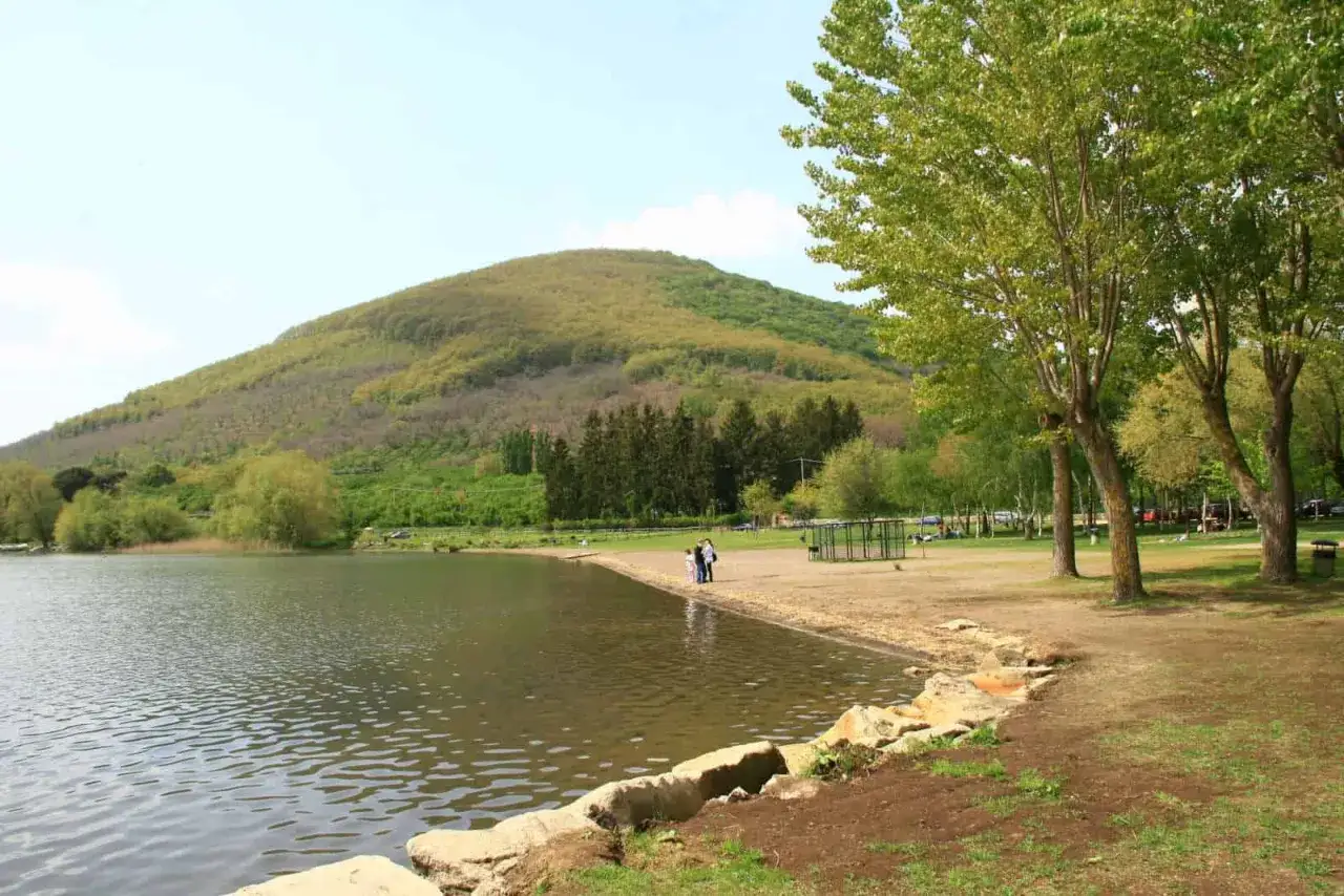 Un tranquillo lago a Caprarola, con una collina verdeggiante sullo sfondo e alberi rigogliosi che ombreggiano la riva sabbiosa.