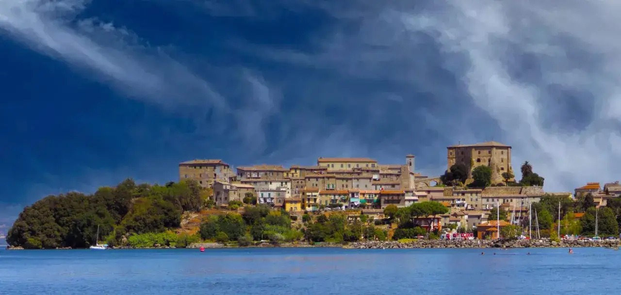 Panoramica di Capodimonte, pittoresco borgo sul lago di Bolsena, con case colorate e un imponente castello sotto un cielo drammatico.